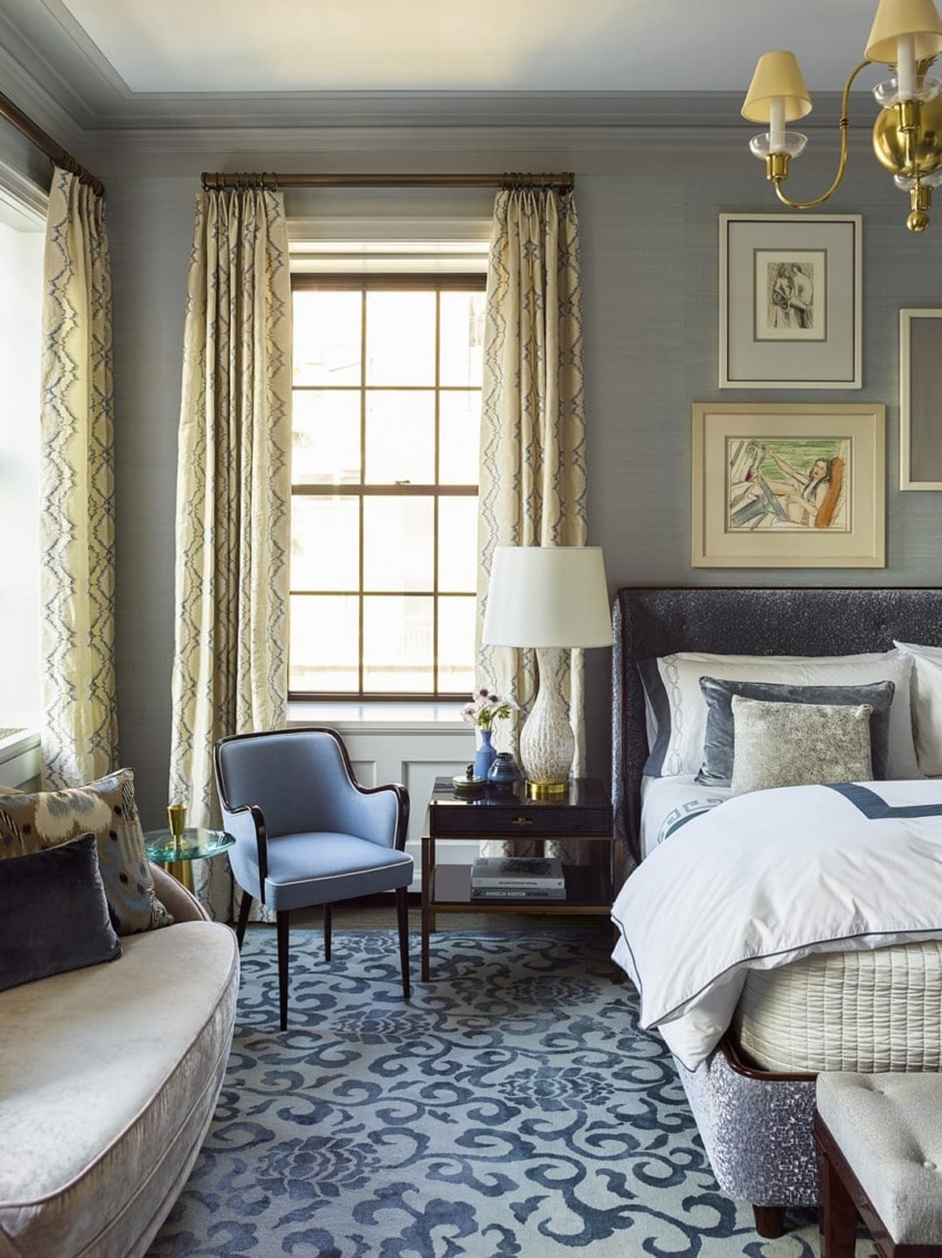 Elegant bedroom interior with patterned rug, cozy bed, plush chair, bedside table, tall windows with curtains, and artwork on walls.