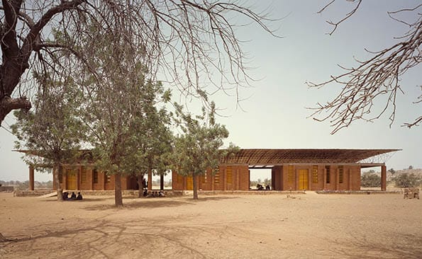 School building in a dry landscape, surrounded by trees, with an open wooden structure and people sitting underneath.