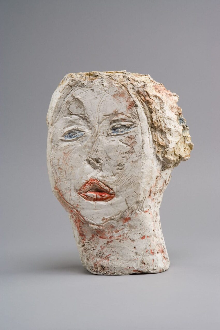 Sculpted clay bust with a textured surface, depicting a face with red lips and a neutral expression against a gray background.