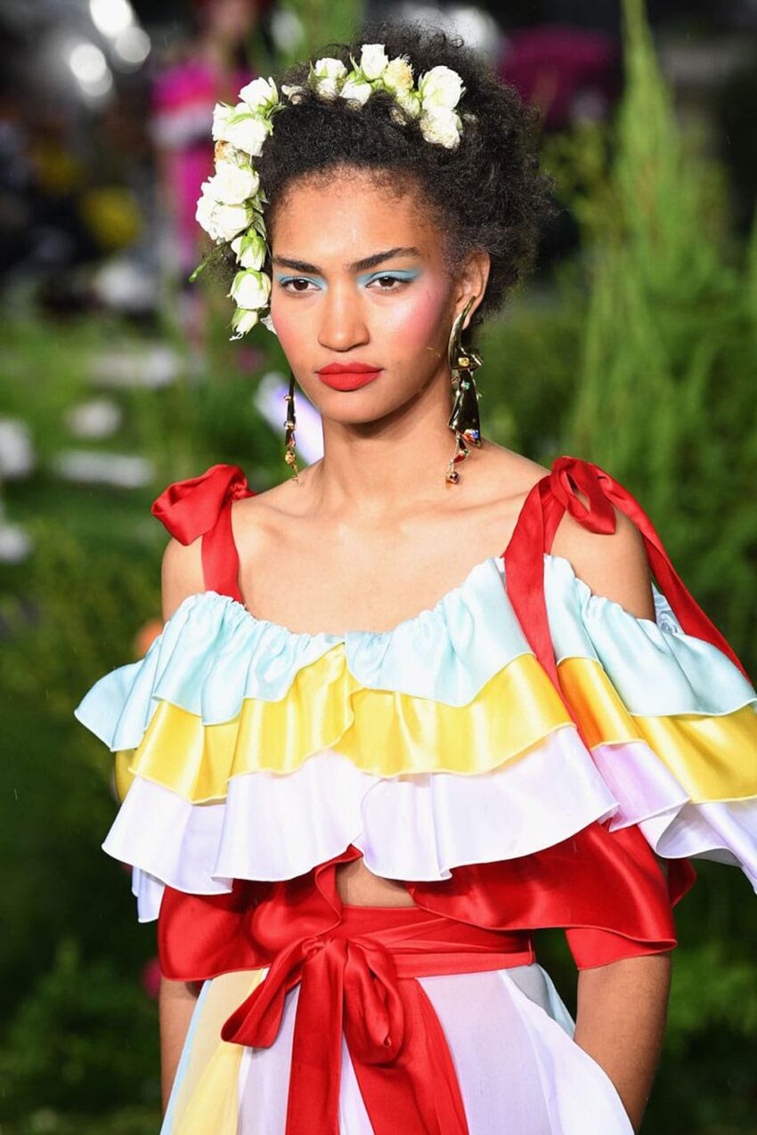 Model wearing a colorful ruffled dress with floral headband on a green outdoor runway.