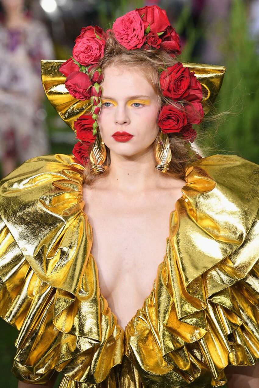 Model with red roses in hair wearing a gold metallic ruffled dress, bold yellow eye makeup, and gold earrings.