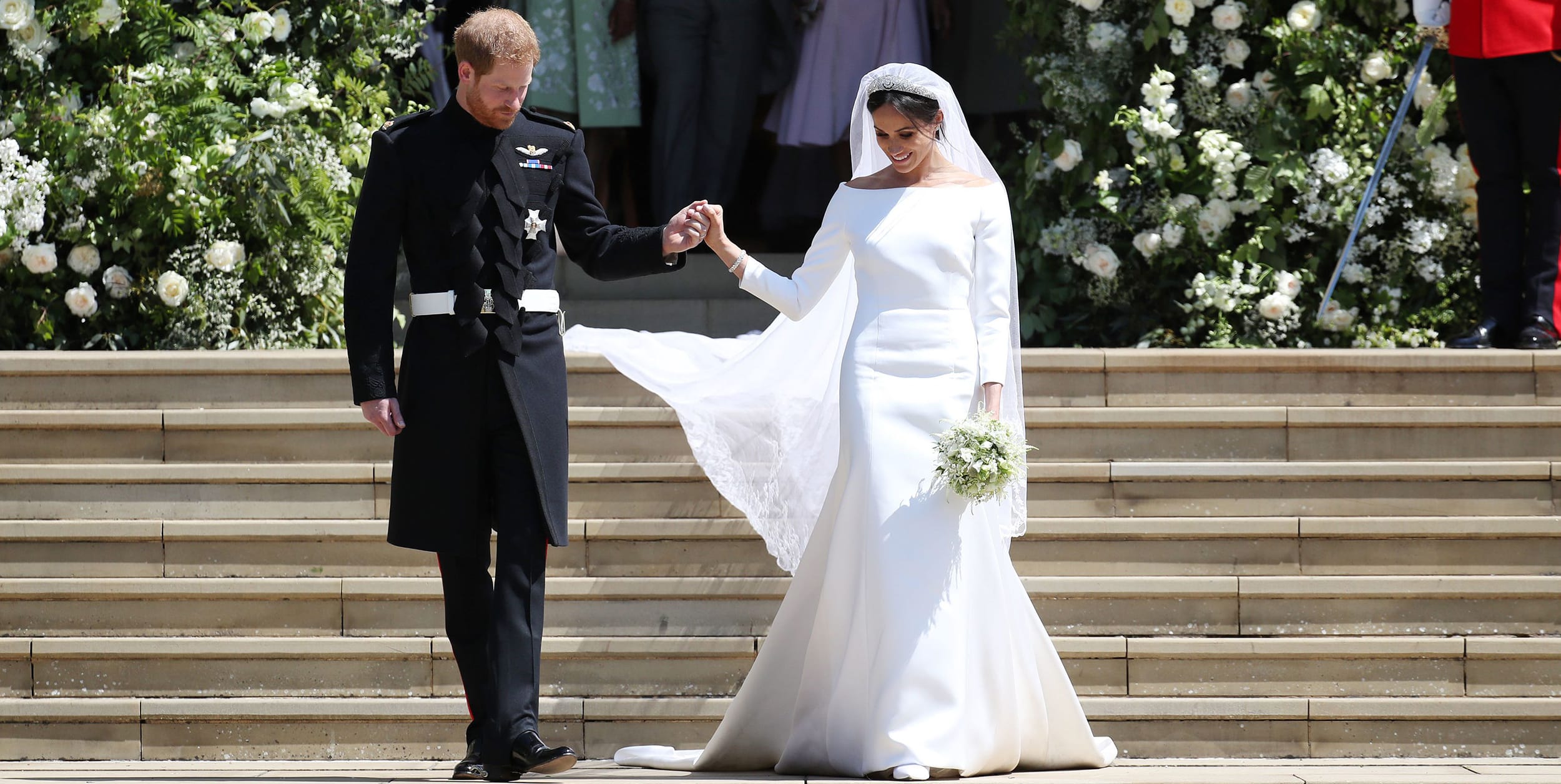 Couple holding hands walking down steps, bride in white gown with veil and bouquet, groom in black suit with military decorations.