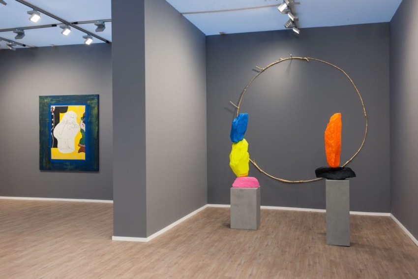 Art gallery with colorful modern sculptures on pedestals and a framed abstract painting on a gray wall.