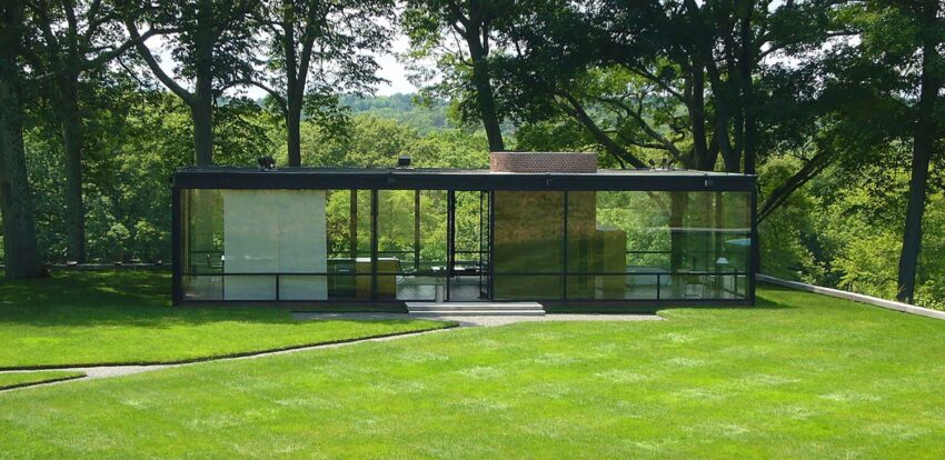 Modern glass house with large windows surrounded by lush green lawn and trees in the background.