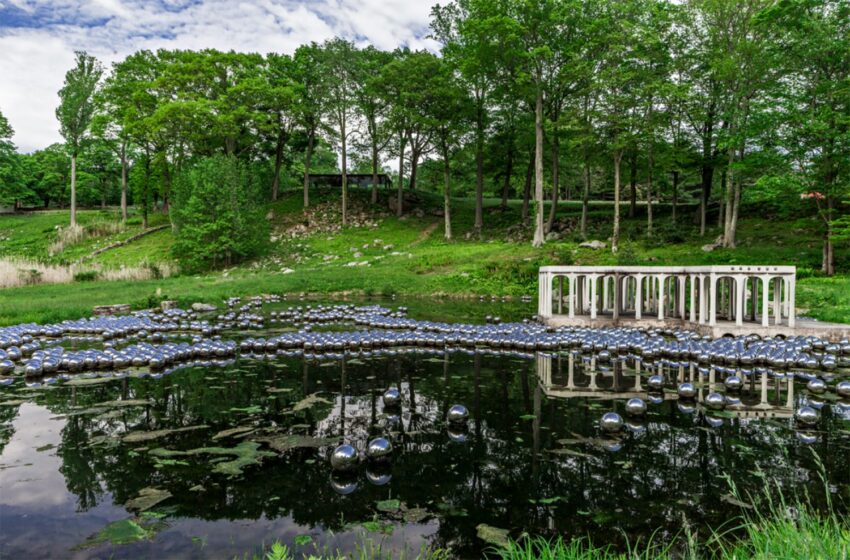 Sculpture installation with silver spheres on a pond surrounded by lush greenery and trees in a park setting.