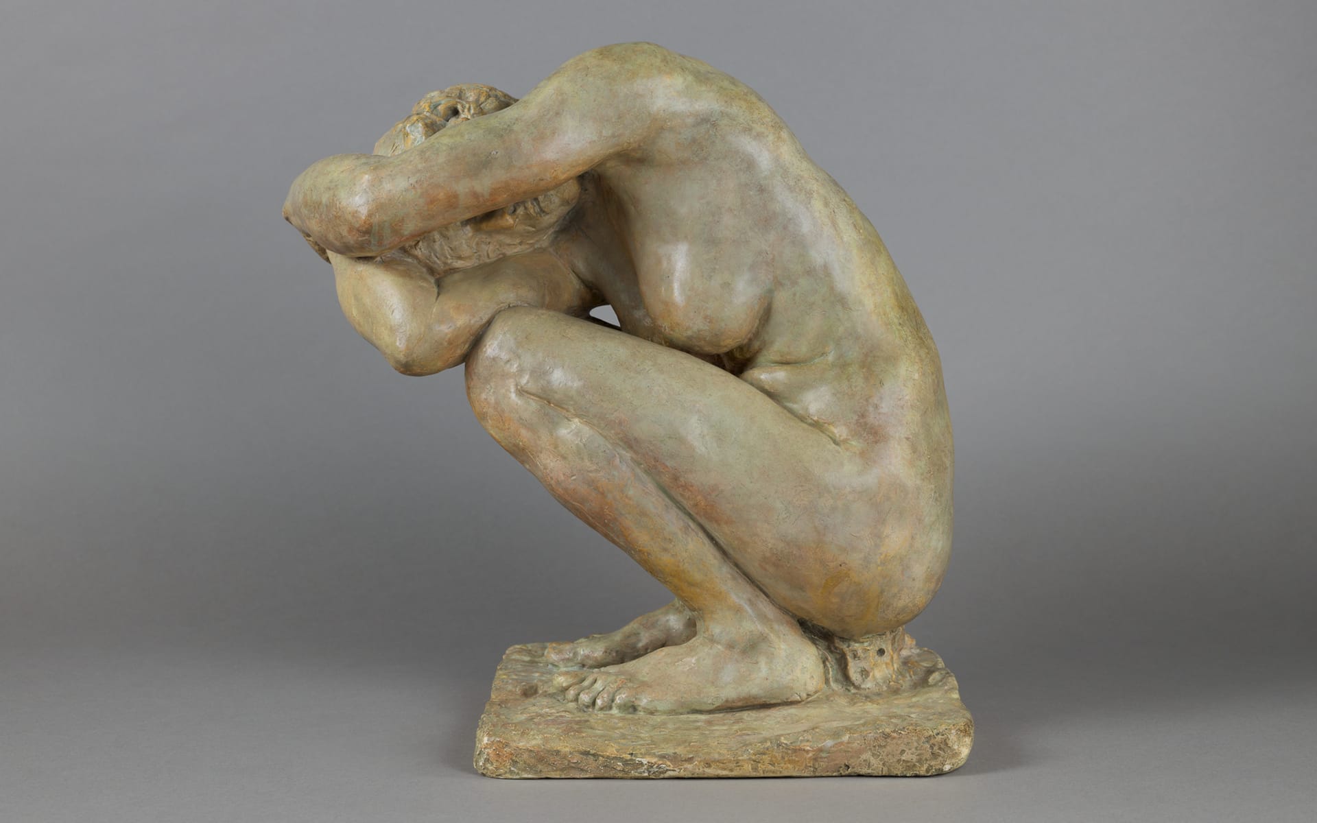Bronze sculpture of a crouching person with arms folded over head on a rectangular base against a gray background.