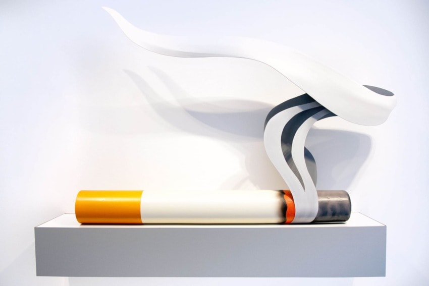 Modern sculpture of a cigarette with abstract white smoke displayed on a minimalist gray shelf against a white background.