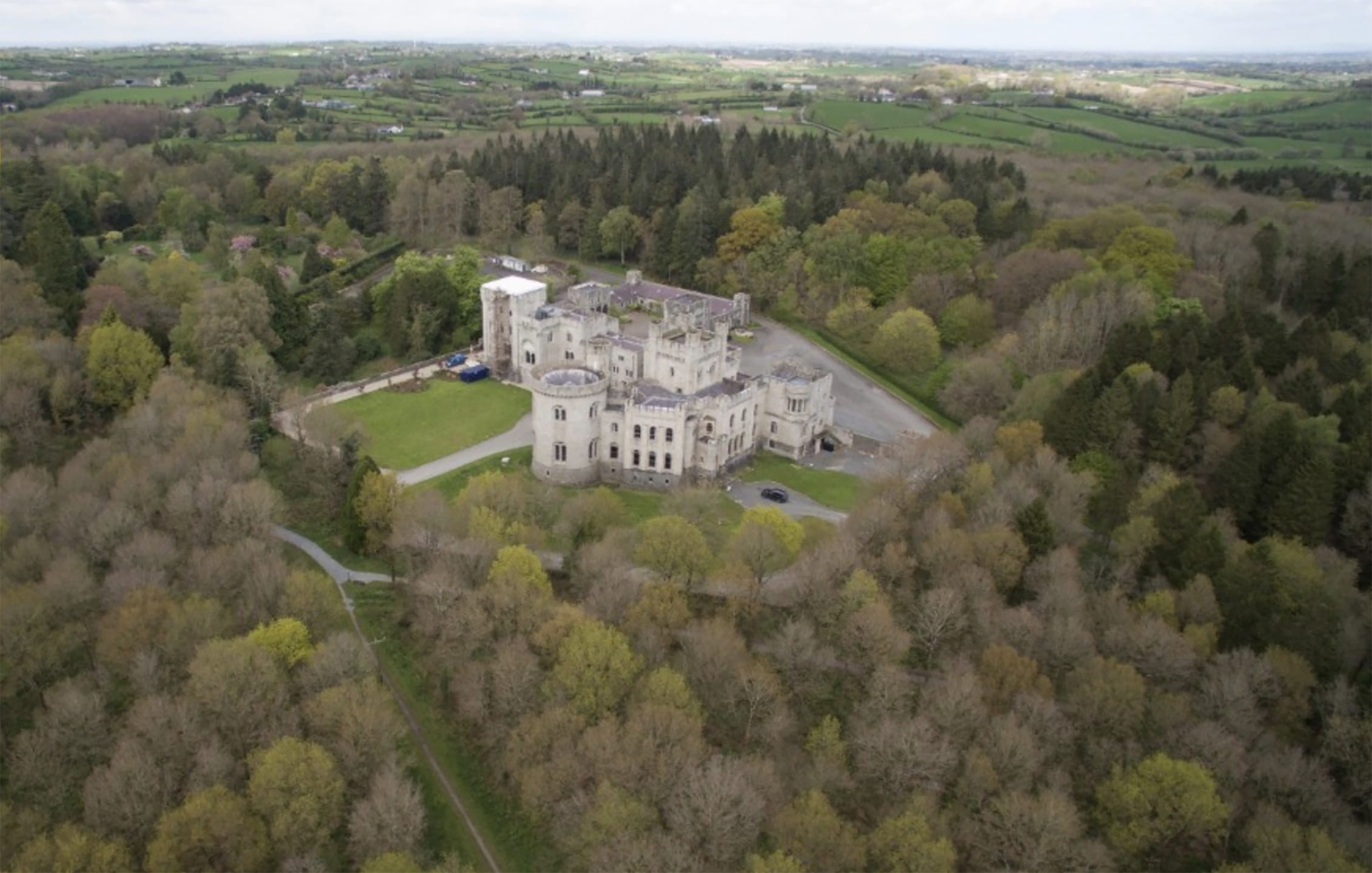 Aerial view of a large, historic castle surrounded by dense woodland and rolling green countryside.