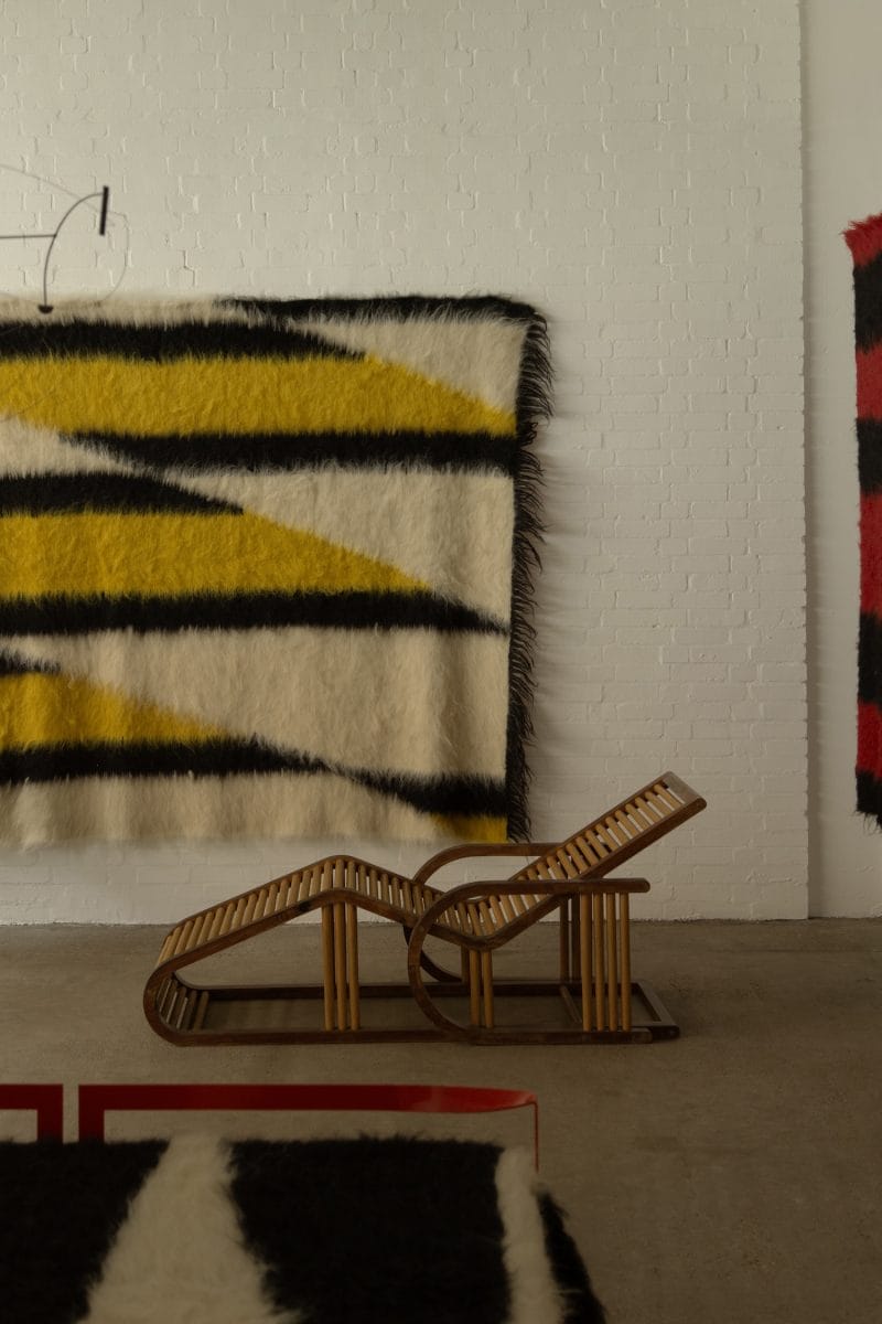 Wooden lounger in front of abstract geometric wall tapestry with yellow, black, and white patterns.