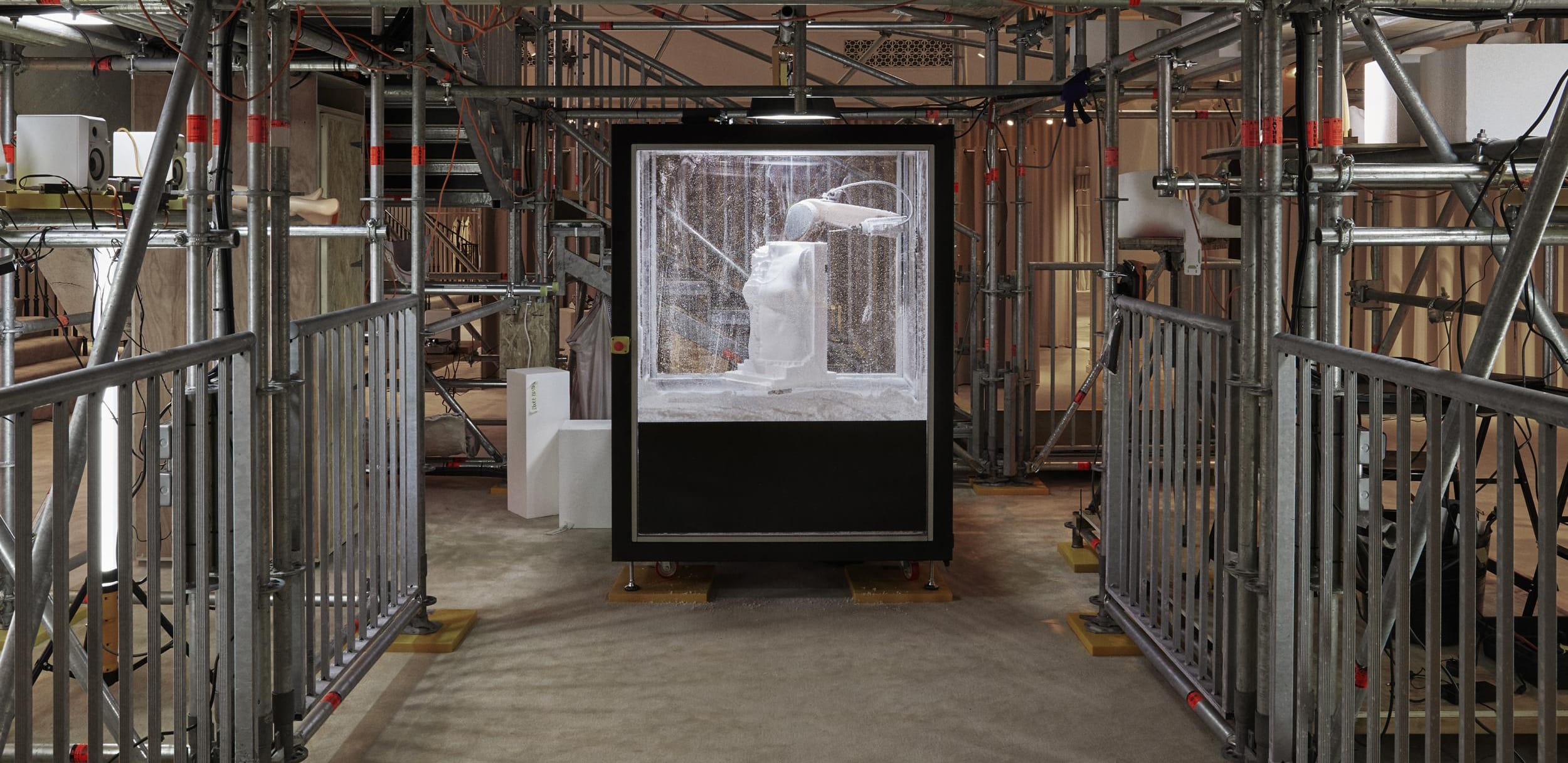 Robotic arm creating a sculpture within a framed cube surrounded by scaffolding in an industrial setting.
