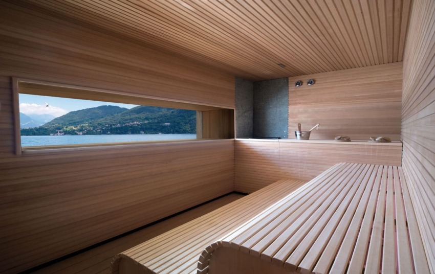 Interior of a wooden sauna with a large window offering a scenic view of a lake and mountains in the background.