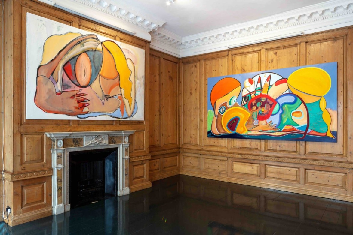 Art gallery interior with two large abstract paintings on wood-paneled walls and a decorative fireplace.