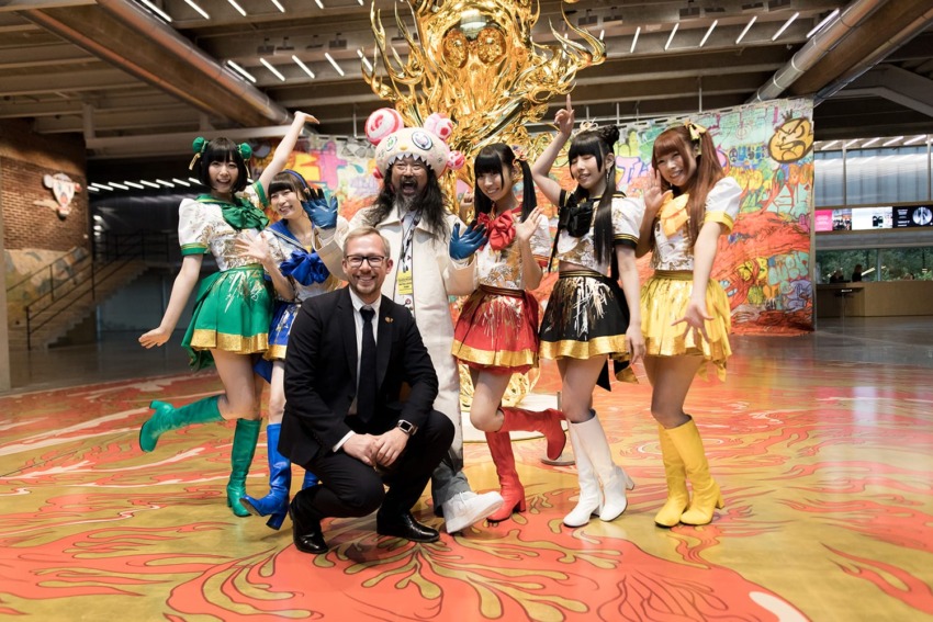 Group in colorful costumes posing with a man in a suit in front of a golden sculpture in an indoor setting.