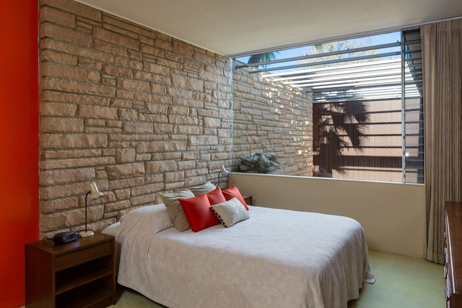 Modern bedroom with stone wall, large window, and minimalist decor featuring white bedding and red accent pillows.