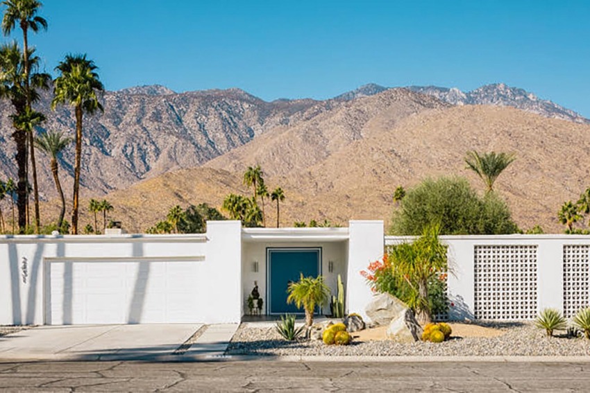 Modern desert home with blue door, palm trees, and mountain backdrop in a sunny landscape.