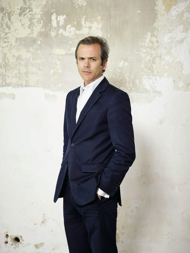 Man in a navy suit standing against a textured wall background.