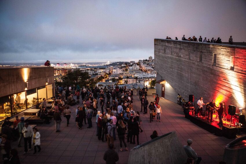 People enjoying an outdoor concert on a rooftop with cityscape views at dusk.
