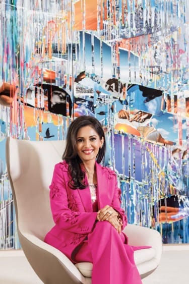 Person in a pink suit sitting on a chair in front of a vibrant abstract artwork with various colorful elements.