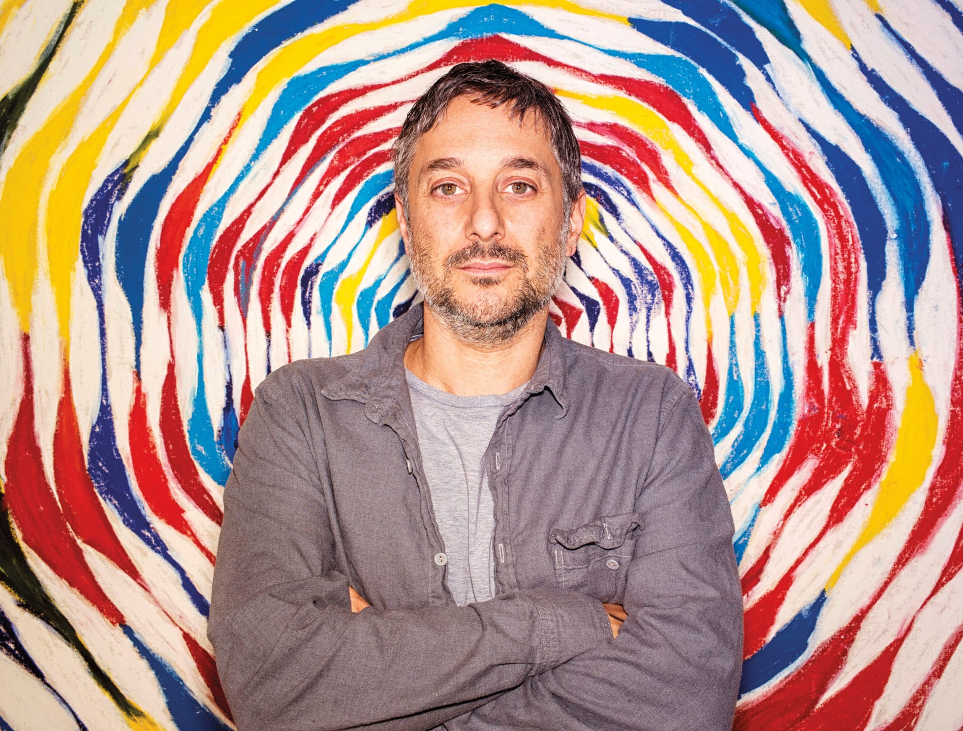 Person standing with arms crossed in front of colorful spiral mural with swirling patterns in red, blue, yellow, and white.