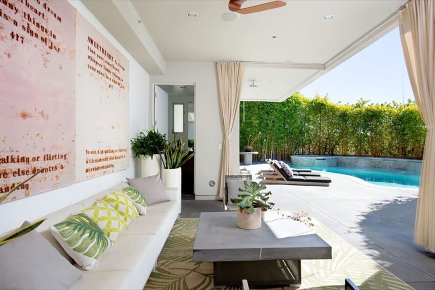 Modern patio with white sofa, green cushions, and plants, opening to poolside view with lounge chairs and greenery.