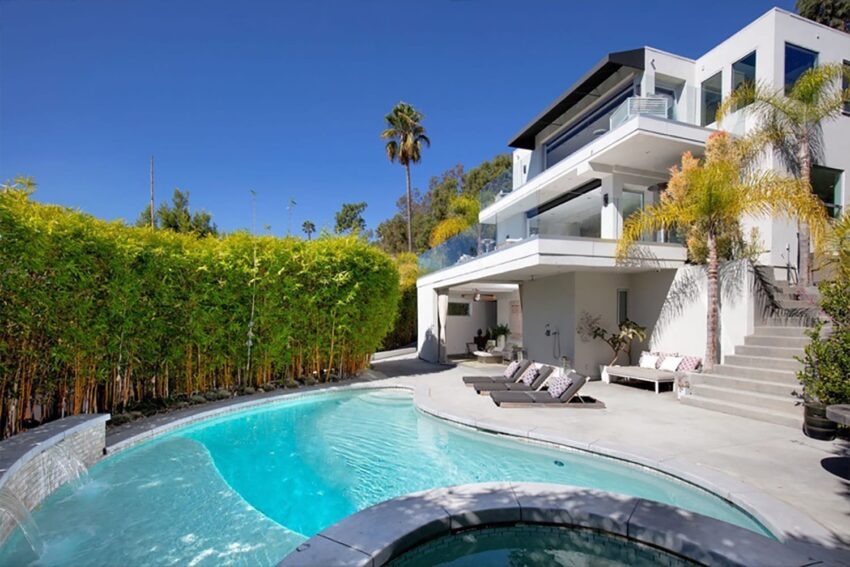 Modern white house with large windows, a pool, and surrounding palm trees under a clear blue sky.