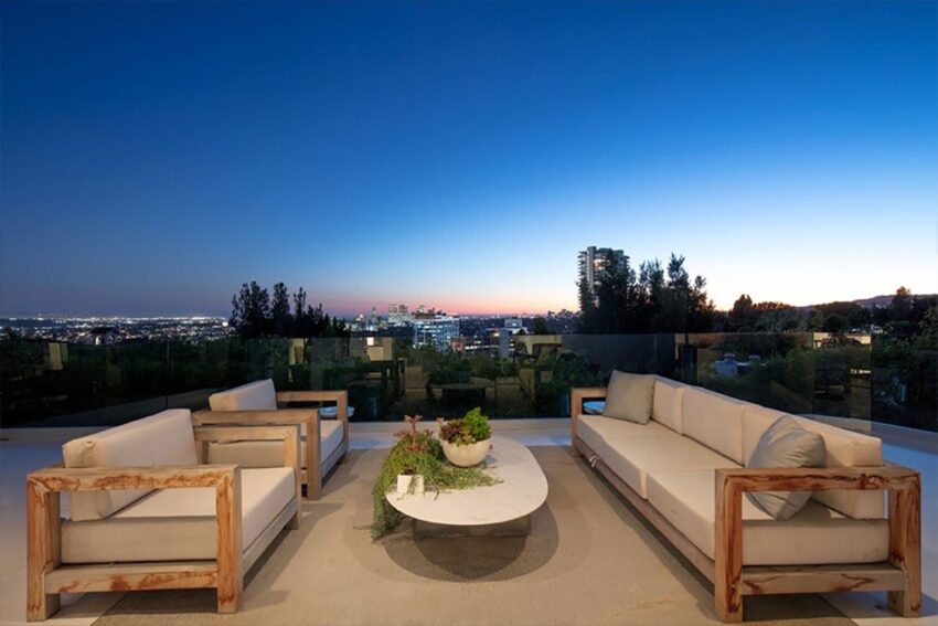 Rooftop terrace at sunset with city skyline view, featuring modern couches and a round coffee table with plants.