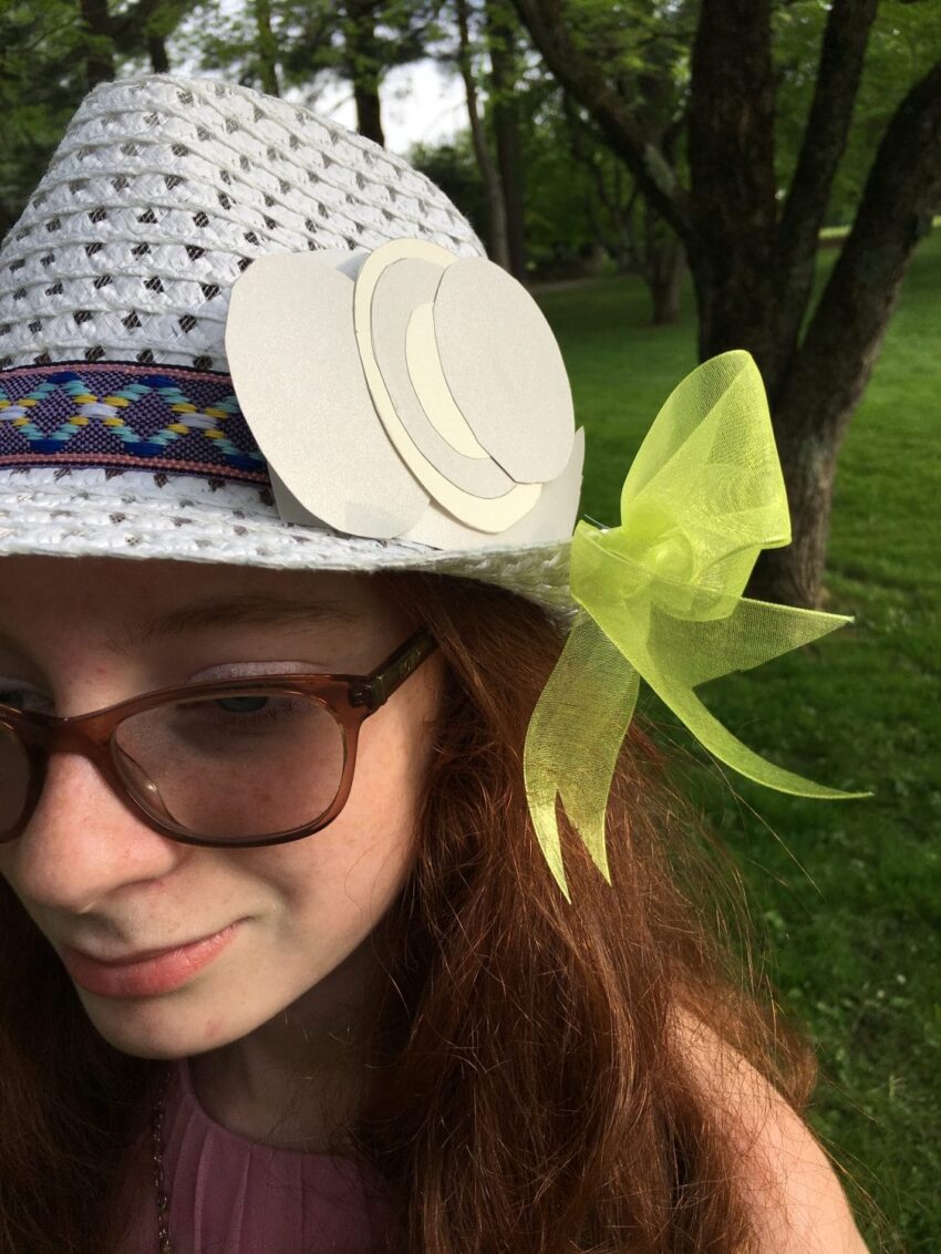 Girl wearing glasses and white hat with a colorful band and yellow bow, standing outdoors in a green park.