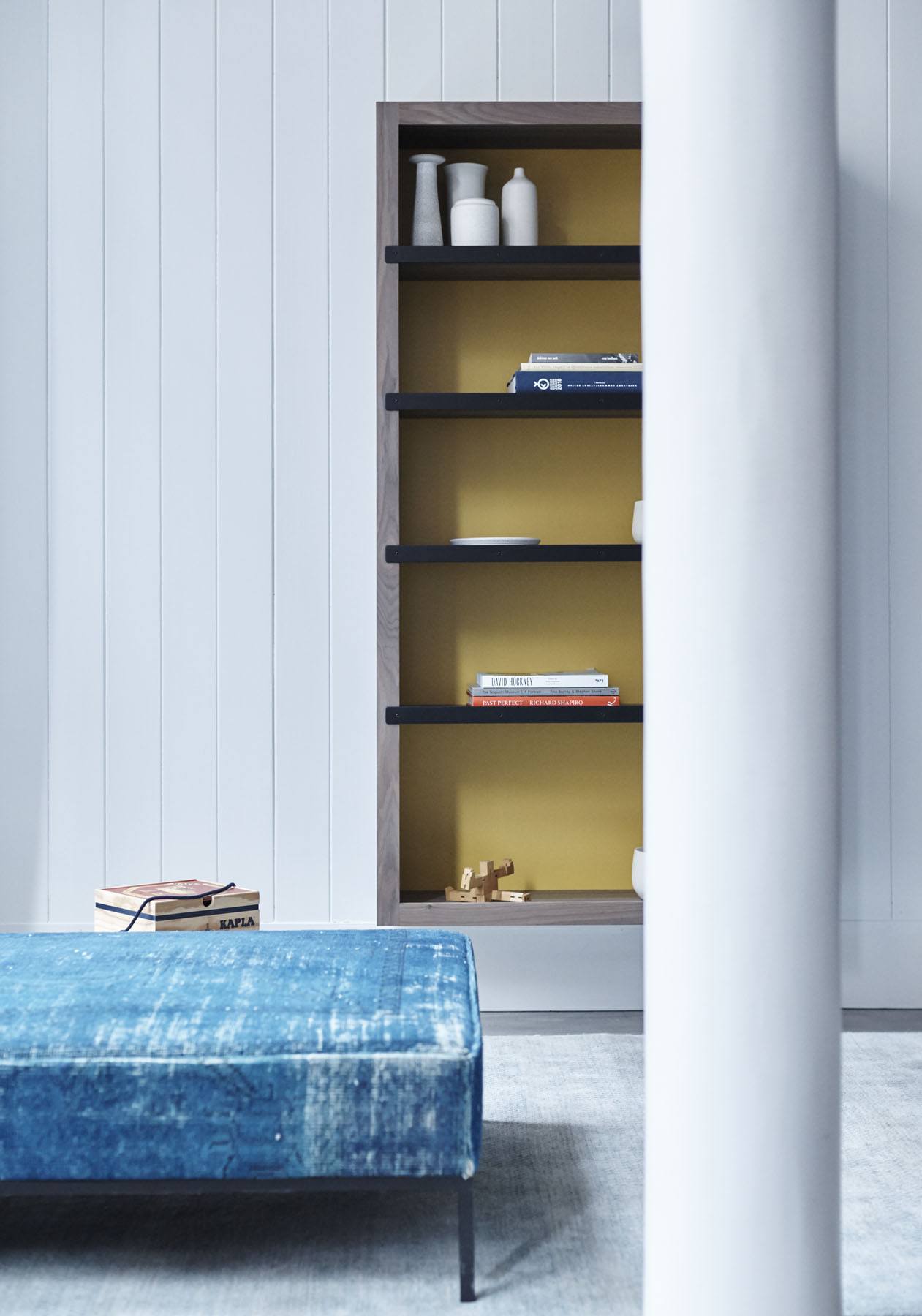 Blue bench, open shelving with books and vases, yellow interior, white paneled wall, and a pillar in a modern room setting.