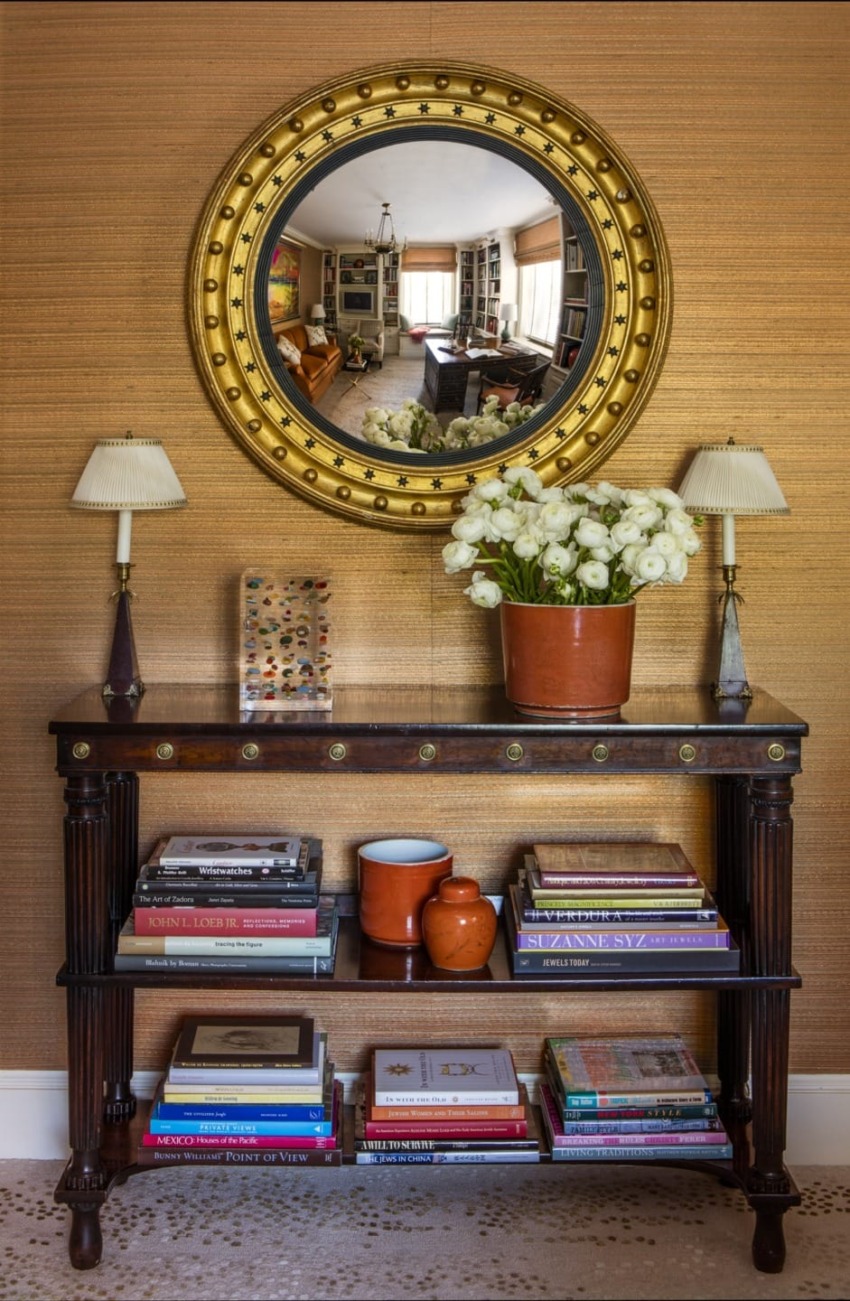 Elegant entryway table with decorative items, round mirror reflecting a cozy living room, lamps, flowers, and books neatly arranged.