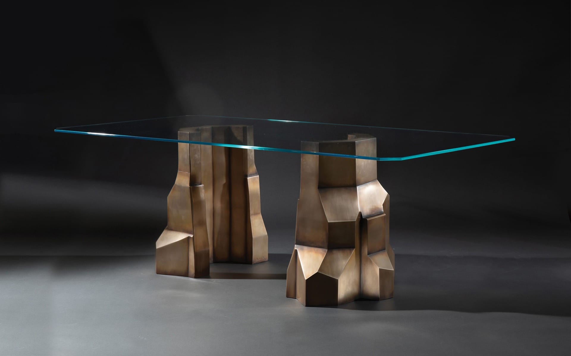 Modern glass-top table with geometric bronze base design on a dark background.