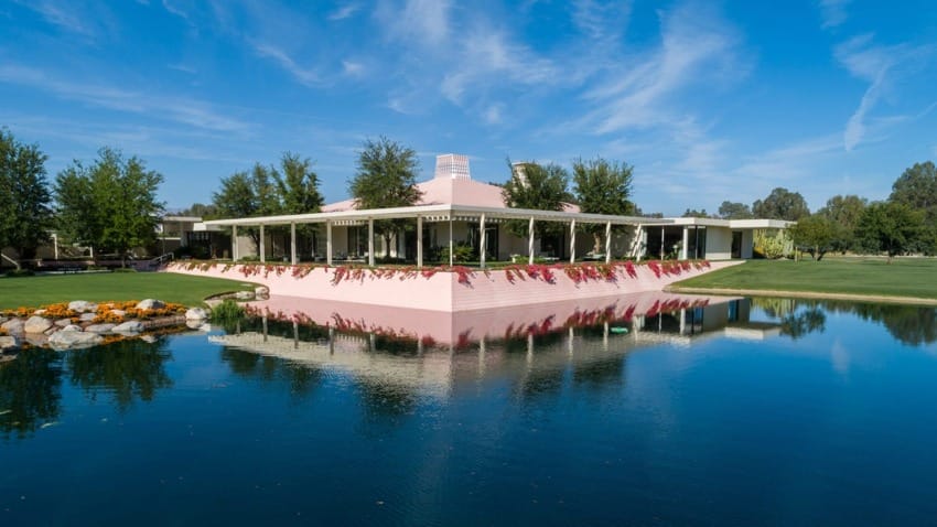 Mid-century modern pink house with large windows surrounded by trees, reflected in a tranquil pond under a blue sky.