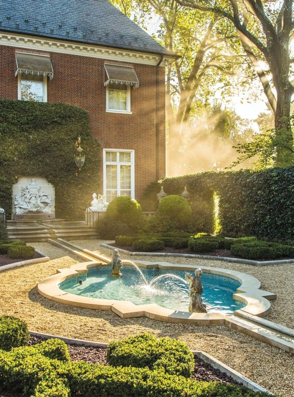 A charming garden with a small fountain, surrounded by hedges and a brick building, bathed in early morning sunlight.