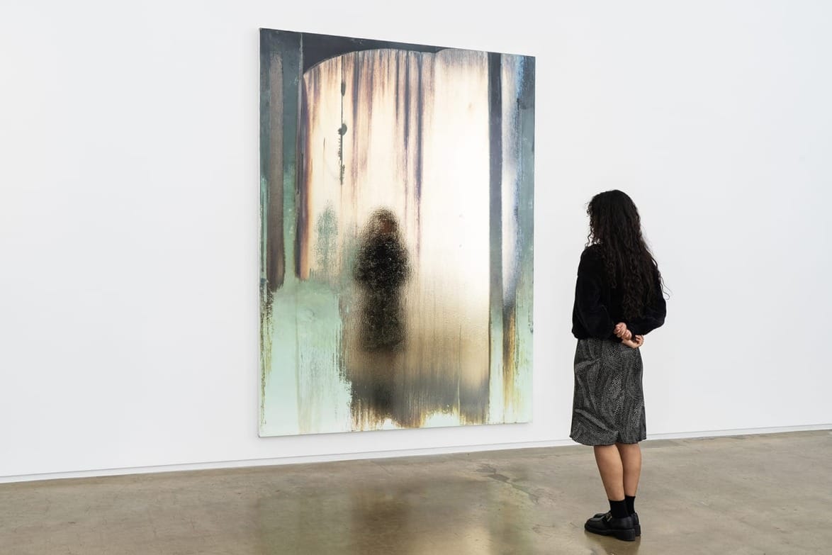 Person observing a large abstract painting with blurred figures and earthy tones in a modern art gallery.