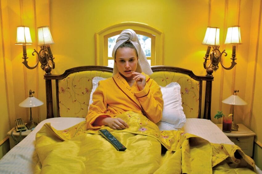 Woman in yellow bathrobe and towel in a yellow room, sitting on bed with remote control, lights on wall sconces.