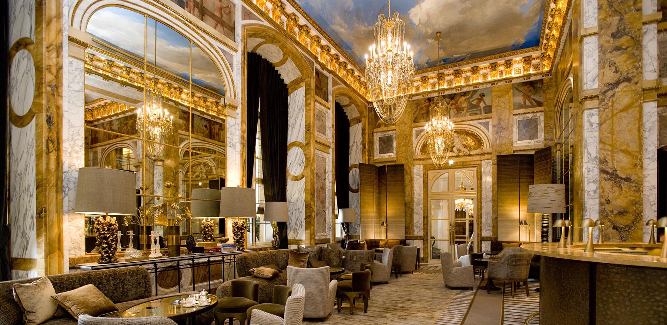 Luxurious hotel lobby with ornate decor, chandeliers, and plush seating under a painted ceiling.