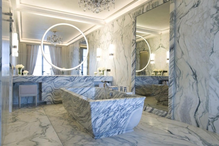 Luxurious marble bathroom with large tub, circular mirror, elegant lighting, and floral decor.