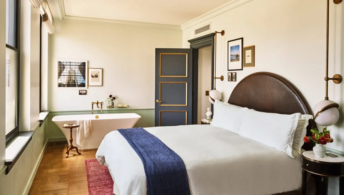 Elegant hotel room with a king-size bed, white linens, blue throw, modern decor, and a freestanding bathtub near large windows.