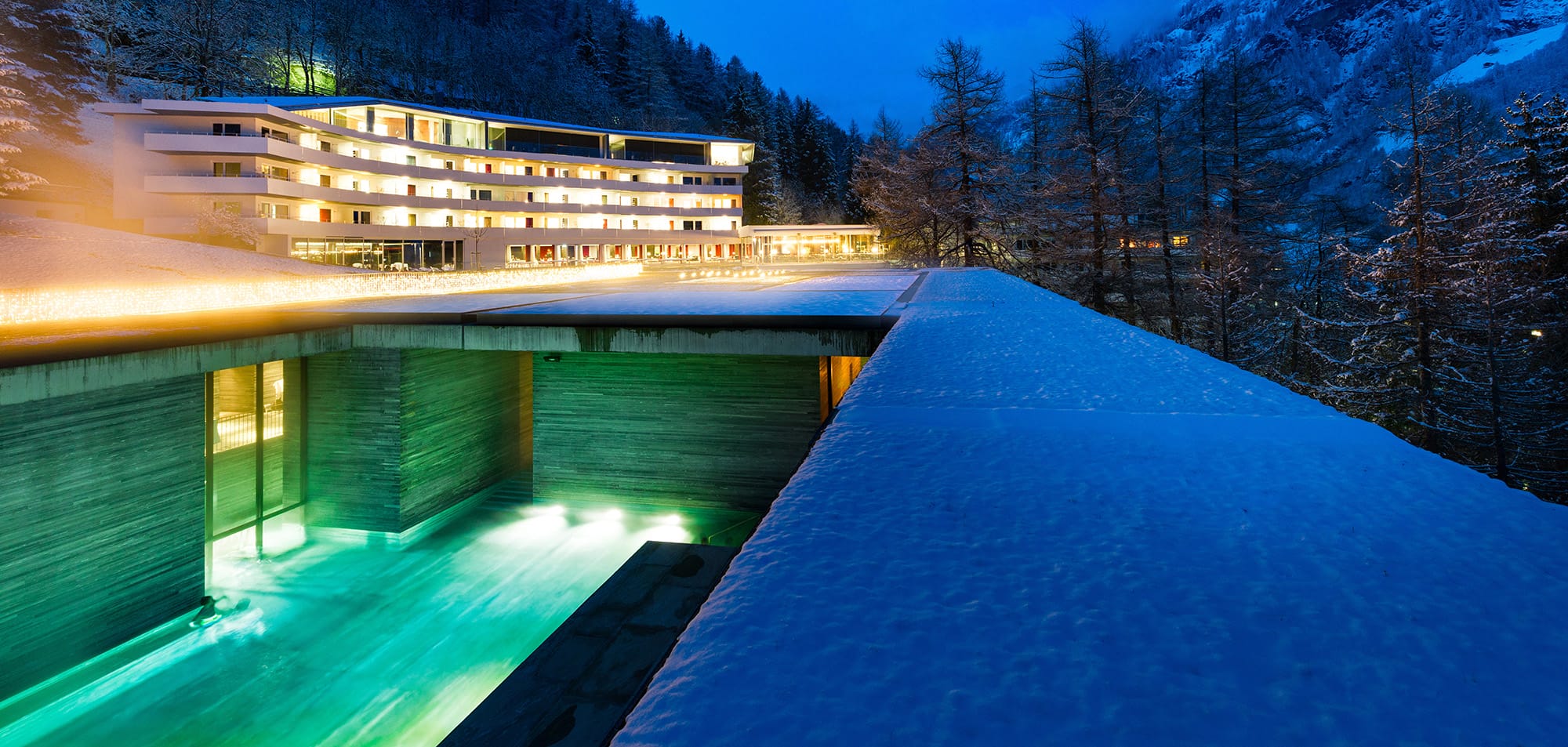 Modern hotel with illuminated windows at night, surrounded by snow-covered landscape and trees, reflecting in turquoise pool.