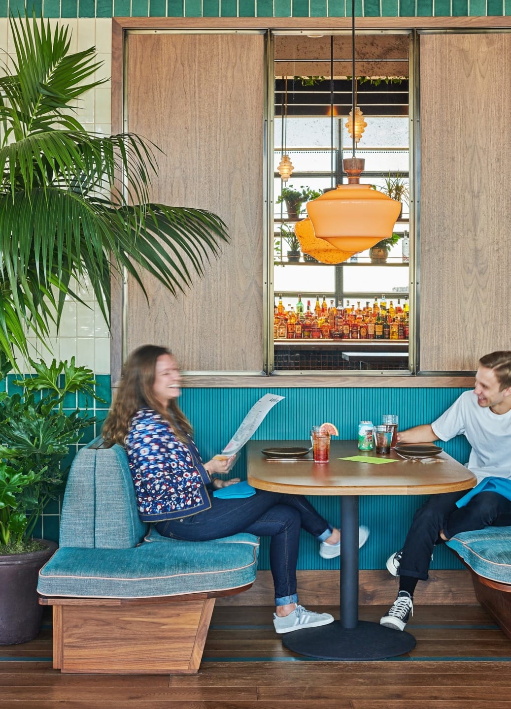 Two people sitting in a vibrant restaurant booth with drinks on the table, wooden paneling, and greenery in the background.