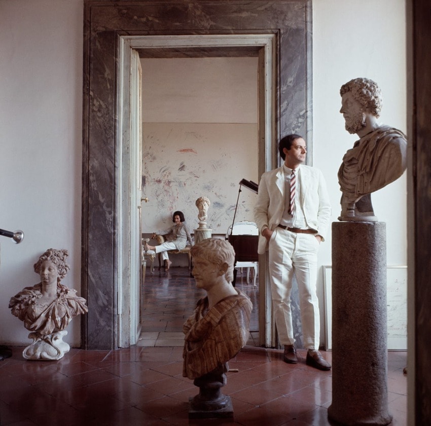 Man in white suit stands among classical sculptures, looking into a room with another person seated in an artist's studio.