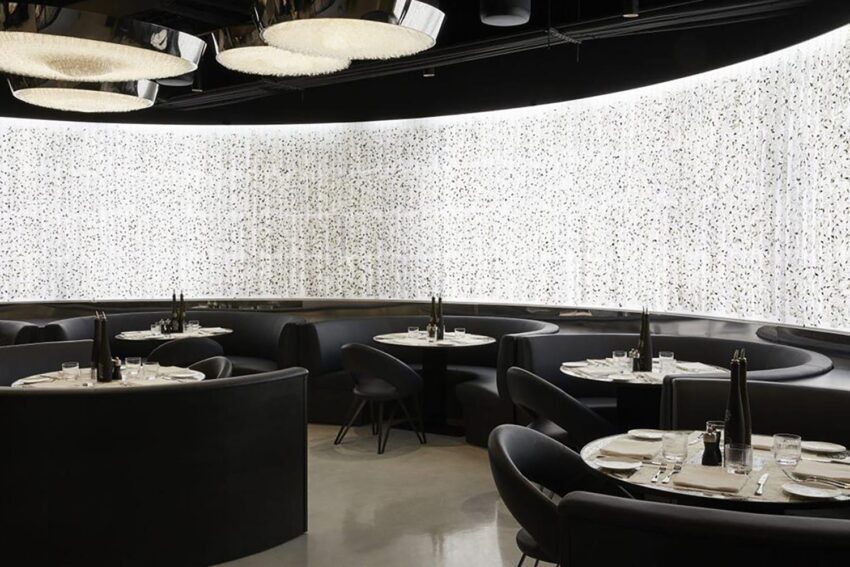 Modern restaurant interior with round booths, elegant black furniture, and soft lighting against a wall of textured white panels.