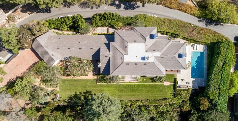 Aerial view of a large house with a gray roof, surrounded by trees, lawn, and a rectangular swimming pool.