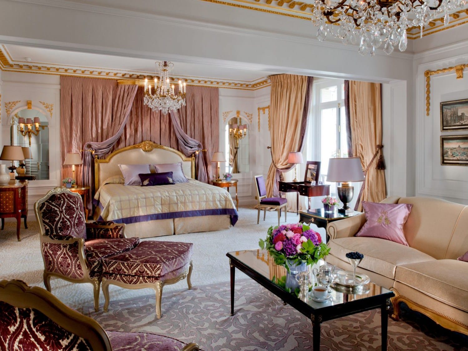 Luxurious hotel suite featuring an elegant bed, ornate furniture, plush seating, chandeliers, and rich drapery.