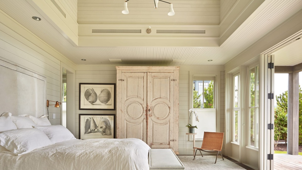 Bright bedroom with white bedding, wooden armoire, artwork on walls, and large windows opening to a patio area.