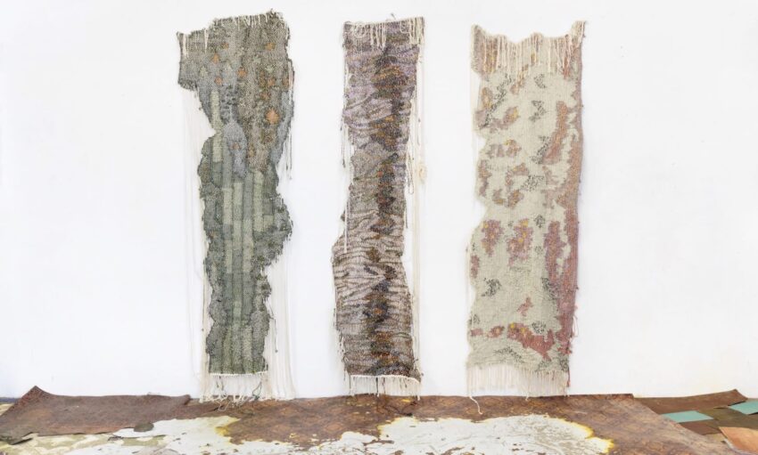 Three large, textured textile art pieces with various colors and patterns, hanging vertically on a white wall.