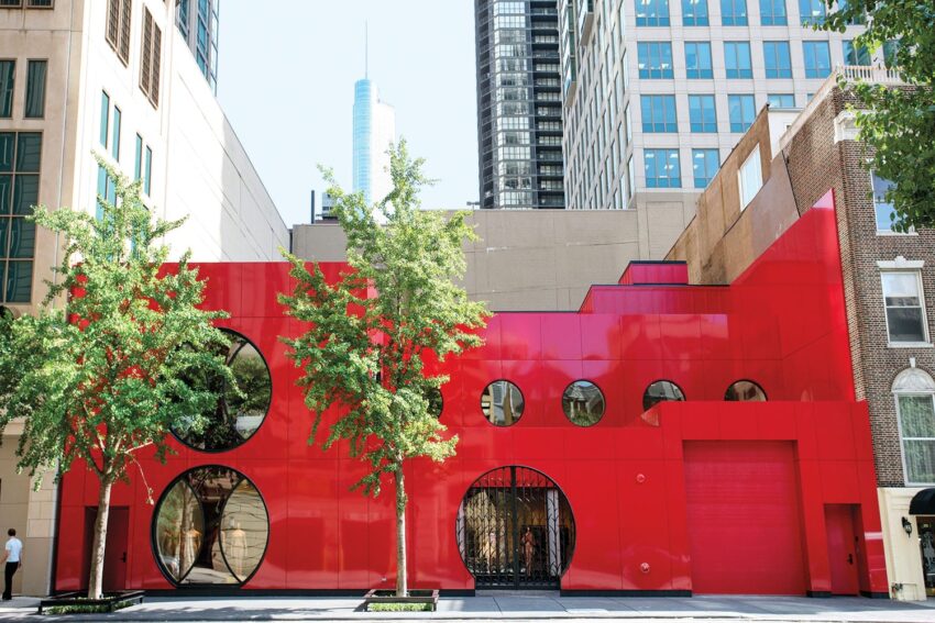 Red modern building with circular windows, surrounded by trees and skyscrapers in the background.
