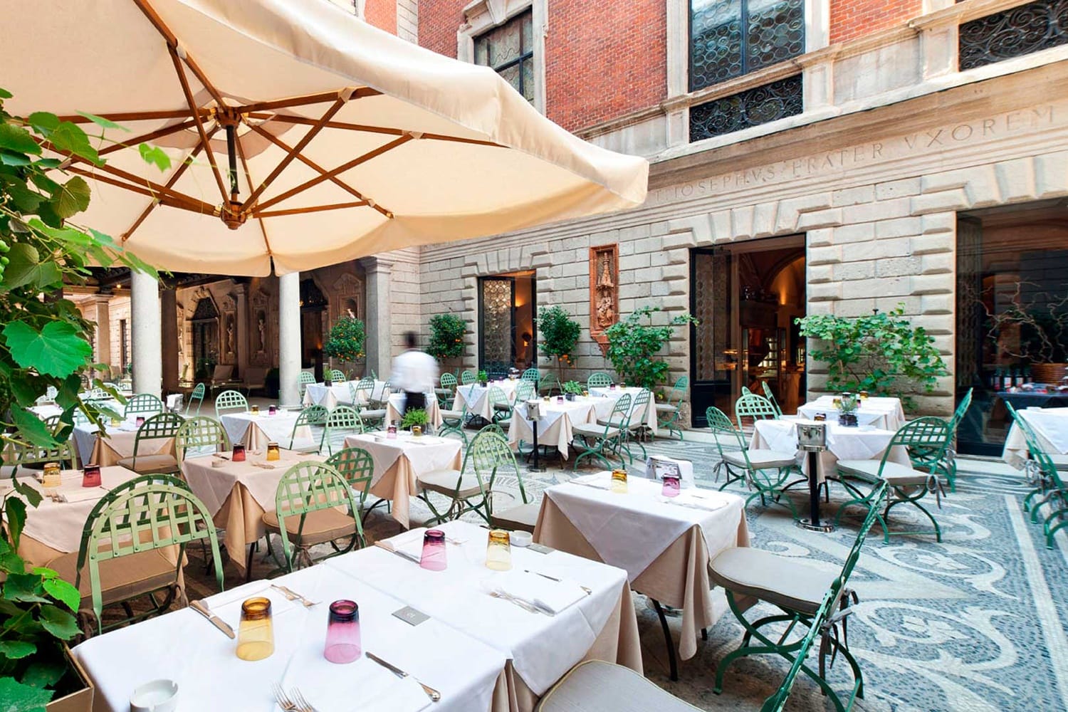 Outdoor restaurant patio with white tablecloths, colorful glassware, and green chairs under a large beige umbrella.