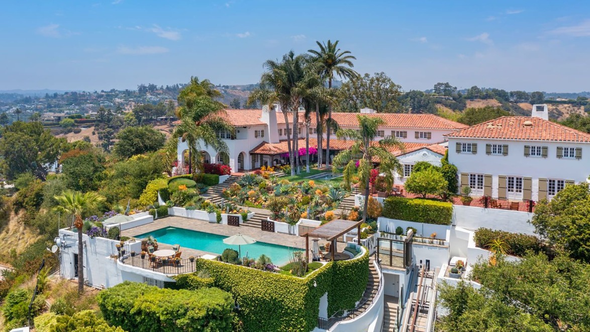 Aerial view of luxurious estate with terracotta roof, surrounded by gardens, palm trees, and a large swimming pool.