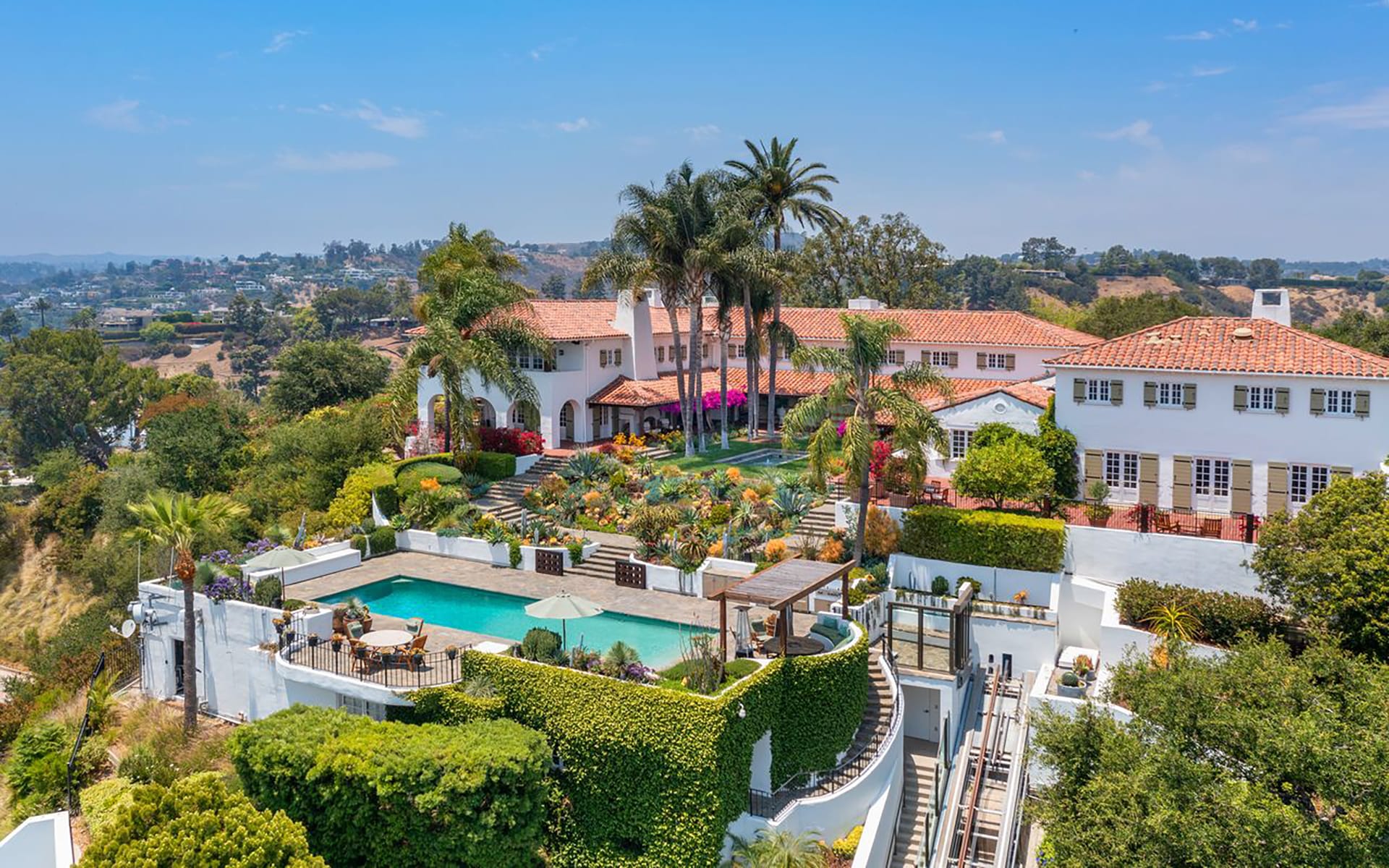 Aerial view of luxurious estate with terracotta roof, surrounded by gardens, palm trees, and a large swimming pool.