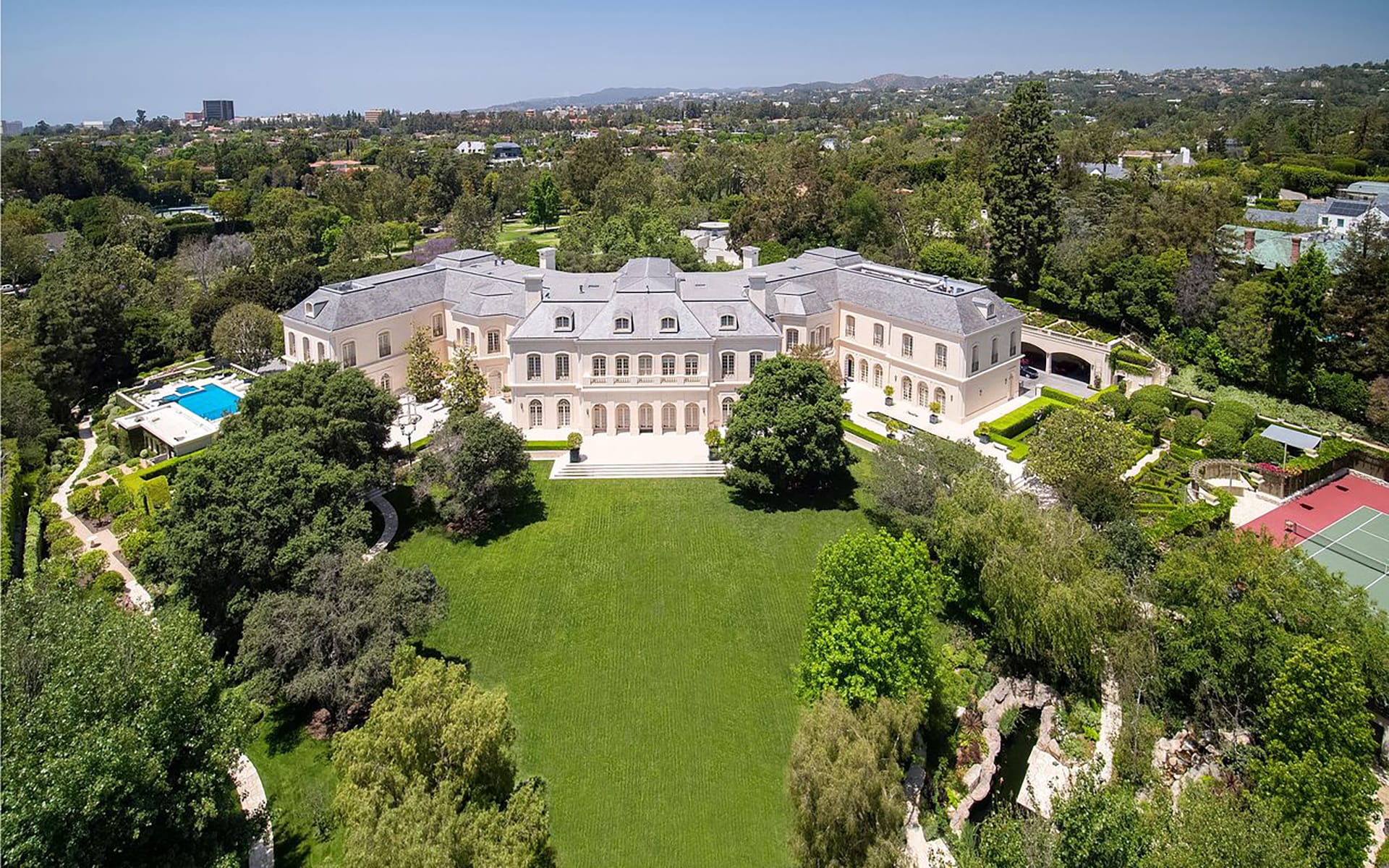 Aerial view of a large mansion with a spacious green lawn, surrounded by trees, a swimming pool, and a tennis court.